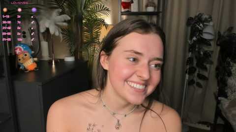 Video of a young woman with fair skin, dark hair, and a smile, wearing a necklace, standing in a dimly lit room with green plants, a stuffed animal, and a shelf with books.