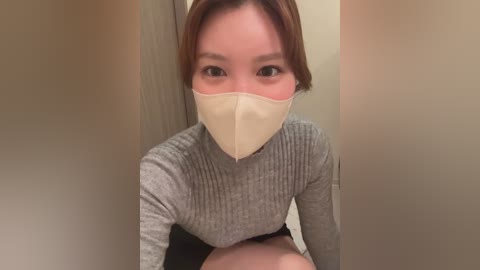 Video of an East Asian woman with fair skin, short brown hair, wearing a gray ribbed long-sleeve top and beige face mask, sitting in a dimly lit room with beige walls.