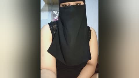 Video of a woman with light brown skin, wearing a black niqab covering her face except for her eyes, a black sleeveless top, and a black headscarf. Background is blurred, with indistinct shapes.