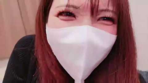 Video of an Asian woman with long, straight red hair, wearing a white face mask, black sweater, and looking slightly upward. Background shows a blurred, beige wall.