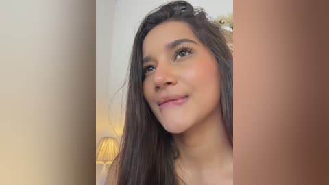 Video of a young woman with medium-brown skin and long, dark hair, smiling slightly. She has a warm complexion and is wearing a simple, light-colored top. The background features a lamp and a blurred, beige wall.