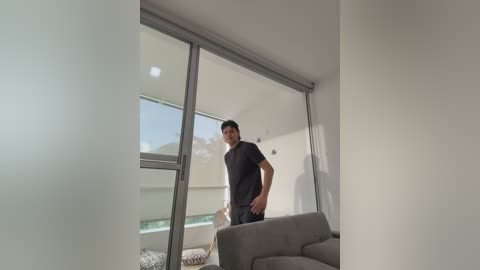 Media: A video of a man with short dark hair, wearing a black t-shirt and jeans, standing in a modern, minimalist living room with a large window and a grey sofa.