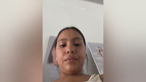Video of a young Latina woman with medium brown skin and straight black hair, wearing a beige top, standing in a bright, white room.