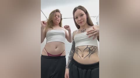 Video of two women in casual attire, one with a white crop top and black skirt, the other in a white crop top and black pants, showcasing tattoos on her arm and lower abdomen.