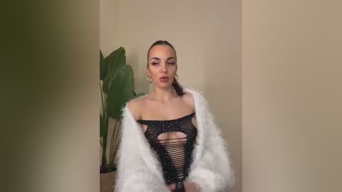 Video of a fair-skinned woman with long, slicked-back hair, wearing a black, revealing, sequin-embellished bodysuit and a white fur jacket, against a beige wall with a large green plant in the background.