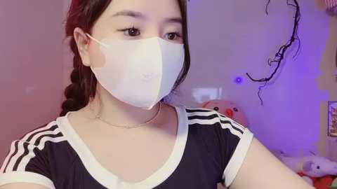 Video of a young woman with light skin, dark hair in braids, wearing a white face mask and black Adidas shirt. Background includes a pink wall, a stuffed animal, and purple lights.