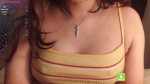 Video of a woman with medium-length brown hair wearing a yellow ribbed tank top with horizontal stripes. She has a silver necklace with a pendant. Background shows a patterned pillow and a green \"Like\" button on the screen.