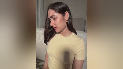 Video of a young woman with medium skin tone, long dark hair, wearing a light yellow t-shirt, sitting on a white bed, eyes closed, smiling, in a softly lit room.