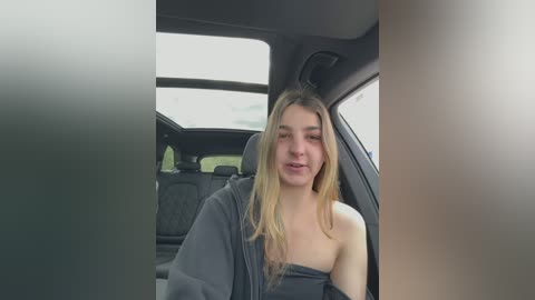 Video of a young woman with long blonde hair, wearing a grey off-shoulder top, seated in a car with grey interior and visible outside sky.