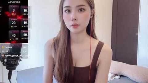 A video of an Asian woman with long brown hair, wearing a brown tank top, sitting in a room with a TV screen displaying weather data.