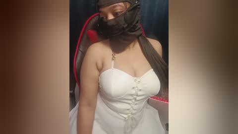Video of a woman in a white dress with a black mask, sitting in a red gaming chair with a black and red headset, in a dimly lit room.