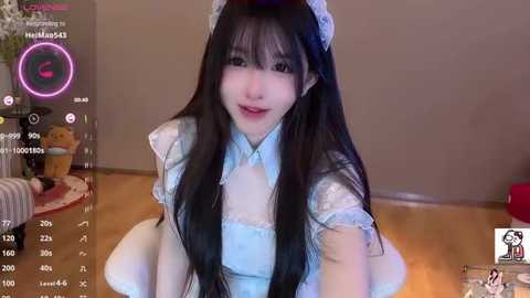 A young Asian woman with long black hair and pale skin, dressed as a maid, sits on a wooden floor. The background shows a TV screen displaying a live stream.