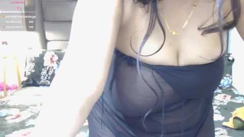 Video of a woman with long dark hair wearing a sheer black dress, showing cleavage and nipples, in a cluttered bedroom with a bed, clothes, and toys in the background.