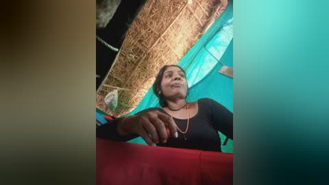 Video of a woman with medium brown skin and dark hair, wearing a black long-sleeve top and gold necklace, sitting under a thatched roof with a green tarpaulin.