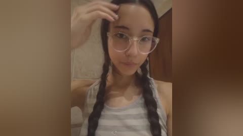 Media: Video of a young woman with long, dark hair in two braids, wearing clear-rimmed glasses and a striped tank top, adjusting her hair in a bathroom with beige tiles and a wooden door.