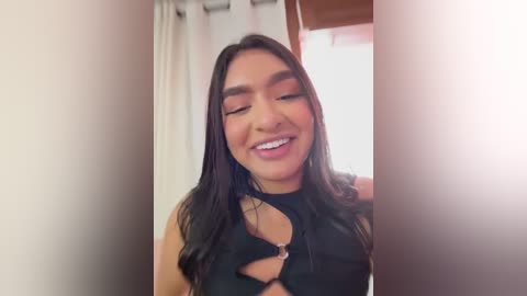 Video of a smiling Latina woman with long, dark hair, wearing a black top with a cut-out design, in a brightly lit room with white curtains and a wooden door.