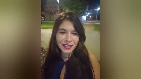 Video of a young woman with long, dark hair and fair skin, wearing a black top, smiling and squinting against the camera. Background shows a blurred outdoor scene with trees and a lit building.