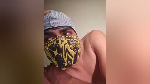 Video of a shirtless, light-skinned person with a grey and white striped headband and a yellow and black patterned mask covering their mouth and nose, against a blurred beige background.