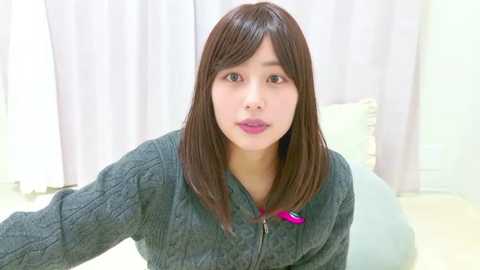 Video of an East Asian woman with straight, shoulder-length dark brown hair, wearing a dark grey, ribbed knit sweater, sitting on a white couch against a backdrop of white curtains and light-colored walls.