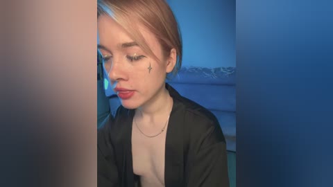 Video of a young, fair-skinned woman with short, blonde hair, wearing a black jacket over a light-colored top. She has a small star tattoo on her cheek and is gazing down with a thoughtful expression. Background features soft blue lighting and a blurred sofa.