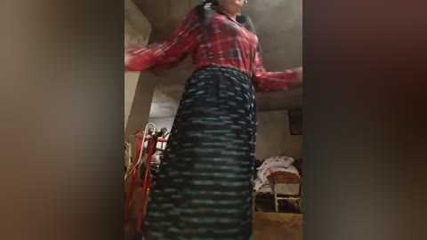 A blurry, low-quality video of a woman in a red plaid shirt and dark, striped skirt, standing in a cluttered room with a red chair and a wicker basket.