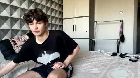 Video of a young Asian man with curly hair, wearing a black T-shirt and shorts, sitting on a bed with a geometric-patterned pillow, surrounded by clothes and a towel rack in a minimalistic bedroom.
