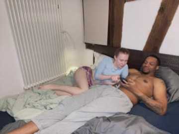 Video of a young white woman with brown hair, wearing a light blue top and pink shorts, lying on a bed next to a shirtless black man with tattoos, both engaged in a playful interaction.