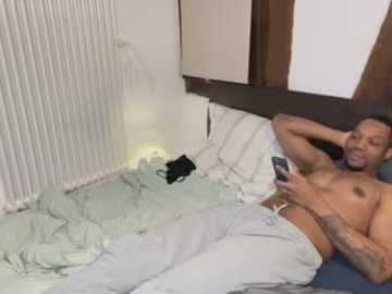 Video of a shirtless, tattooed, muscular Black man with a phone in bed, white sheets, and a black cat on the left.