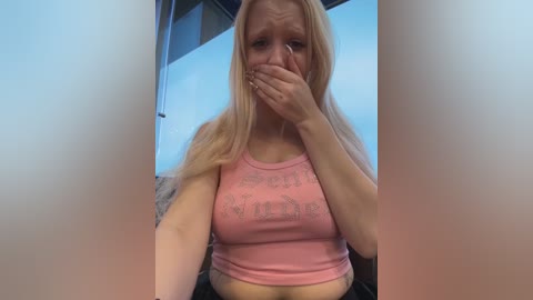 Video of a blonde woman with fair skin, wearing a pink tank top, covering her mouth, seated indoors, with blue walls and a bed visible.