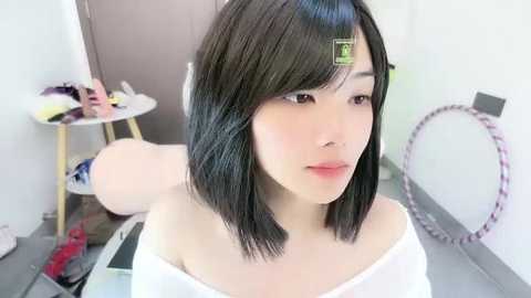 Video of an Asian woman with straight black hair, light skin, and pink lipstick, wearing a white off-shoulder top, standing in a brightly lit room with a small table, chair, and circular mirror.
