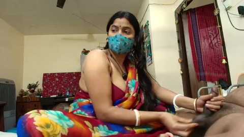 Video of a South Asian woman in a colorful saree, with blue mask, sitting on a bed in a modest room, using a syringe on a man's penis.