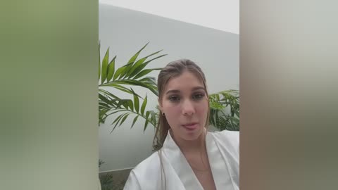 Video of a young woman with fair skin, brown hair pulled back, wearing a white robe, standing in front of a light gray wall with lush green potted plants in the background.