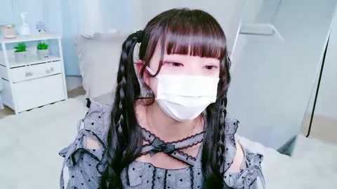 Video of an Asian woman with long, black pigtails, wearing a grey, polka-dotted dress with a lace collar and white face mask, in a modern, light-colored room with white furniture and green plants.