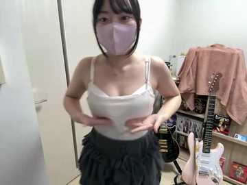 Video of an Asian woman with a slender build, wearing a white tank top and black ruffled skirt, face masked, standing in a cluttered room with shelves of DVDs and a guitar.