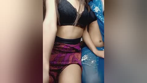 Video of a young woman with light brown skin, wearing a black lace bra and high-waisted, glittery purple shorts, sitting on a blue plastic chair with a floral pattern.