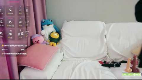 Video of a messy white couch with a blue frog plushie, a pink pillow, and scattered toys, partially covered by a calendar featuring a pink background and numbers.