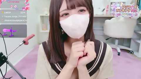 A video of an East Asian woman with straight brown hair and a white face mask, kneeling on a pink carpet, hands clasped, wearing a sailor-style school uniform, in a bright, modern room.