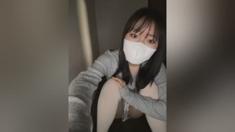 Video of a young Asian woman with long black hair, wearing a white mask, gray hoodie, and white socks, crouching in a dimly lit hallway.