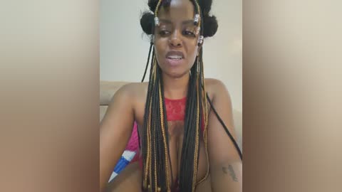 Video of a young Black woman with braided hair and hoop earrings, wearing a red top, sitting indoors.