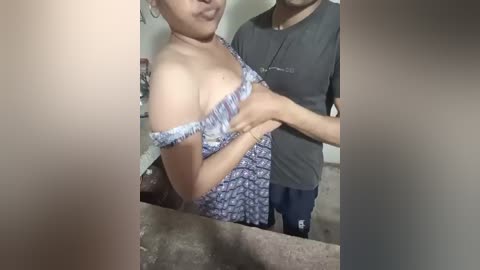 Video of a woman in a floral dress with a man, blurred, in a bathroom with a sink and mirror in the background.