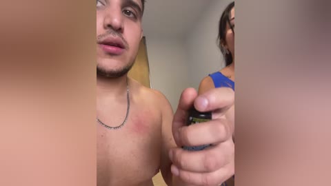 A video of a shirtless man with a light brown complexion and short dark hair, wearing a silver necklace, applying sunscreen to his upper chest. A woman with dark hair and a blue tank top is partially visible in the background.
