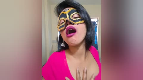 A video of a woman with medium skin tone, wearing a flashy black and gold masquerade mask, bright pink lipstick, and a vibrant pink top. She's indoors, against a blurred background.