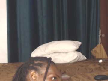 Video of a woman with dark skin and braided hair, lying on a bed with a beige patterned duvet. She's partially visible, with a white pillow behind her head. The background features dark teal curtains.