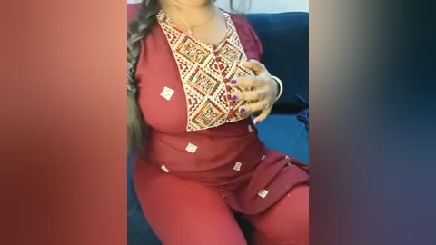 A video of a South Asian woman in a red salwar kameez with intricate embroidery, showcasing her large breasts and curvy figure, posing in front of a dark blue sofa.