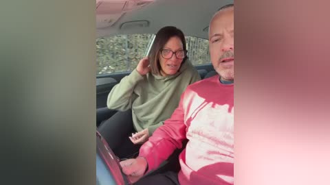 A candid video shows a woman with short dark hair, wearing glasses and a green sweater, gesturing excitedly. A man with a gray beard, balding, and a red shirt is driving. They are inside a car with a sunlit view of greenery outside.