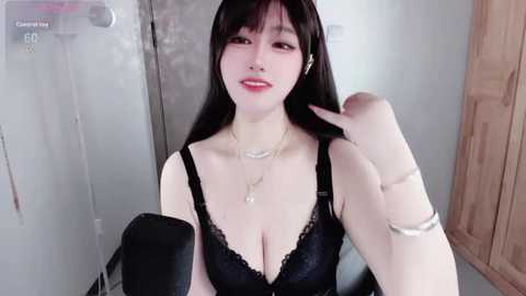 A video of an East Asian woman with long black hair and fair skin, wearing a black lace bra, standing in a bathroom. She has a nose ring and a silver bracelet on her left wrist.