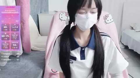 Video of an Asian woman with long black pigtails wearing a white Hello Kitty shirt, face mask, and pink chair. Background shows a pink wall and bed.