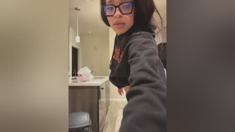 Video of a young Asian woman with black hair and glasses, wearing a black hoodie, taking a selfie in a small, dimly lit room with beige walls, a desk, and a trash can.