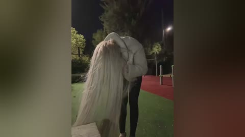 A video of a bride in a long, flowing white veil and a groom in a dark suit, kissing passionately under a tree at night, with a red soccer field in the background.