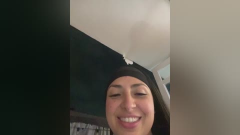 A video of a smiling woman with light brown skin and dark hair, wearing a black headband, captured indoors. The background features a white ceiling with a visible crack and dark, indistinct surroundings.